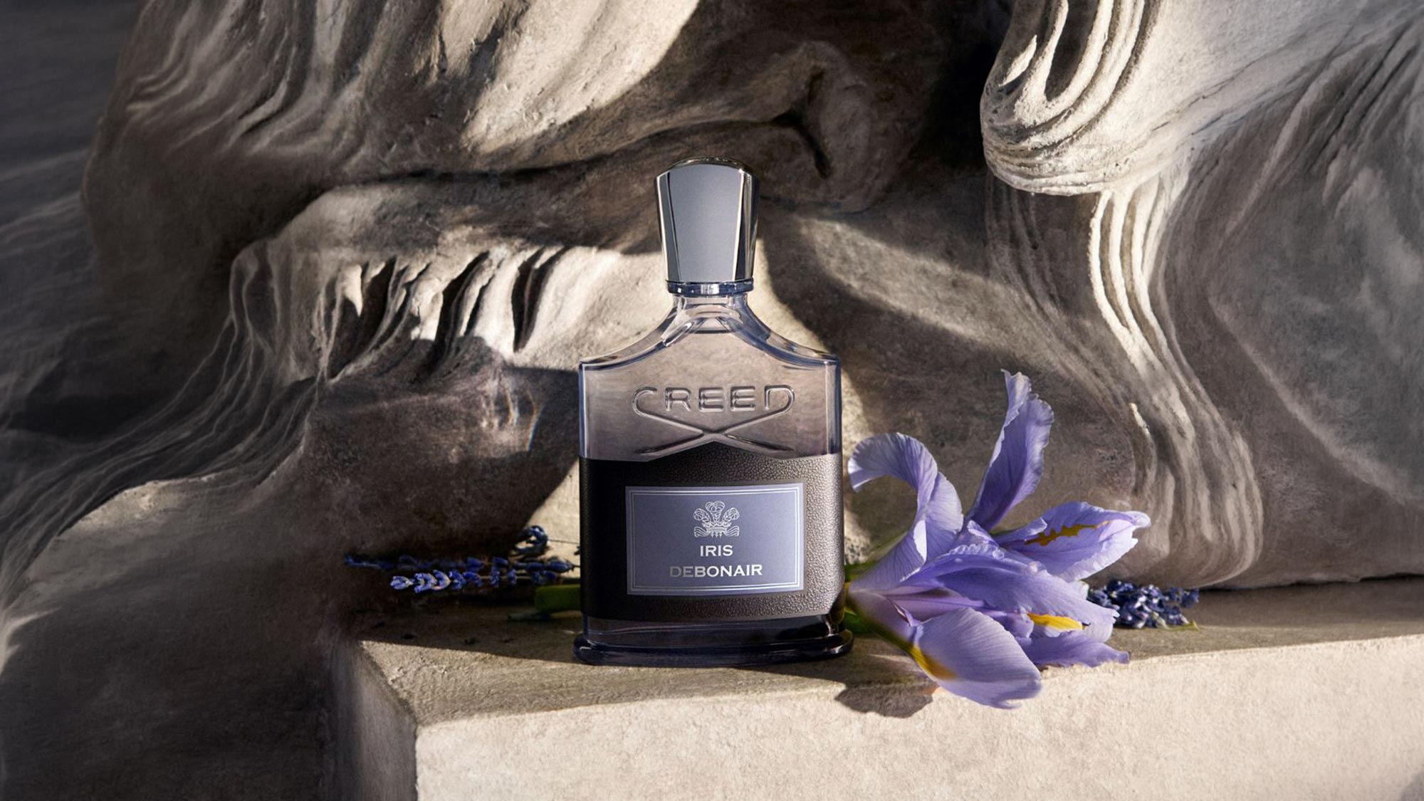Creed Iris Debonair Eau de Parfum bottle with purple iris flowers and lavender accents on stone background