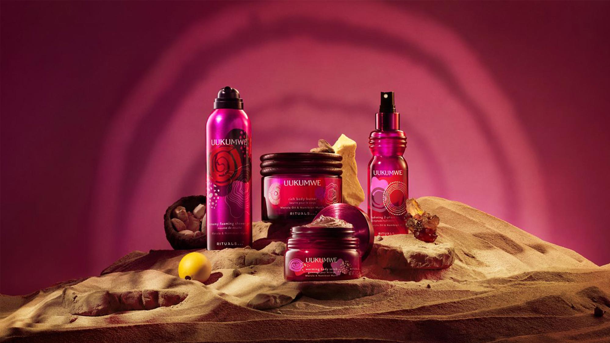 Uukumwe Rituals collection displayed on sand with pink background, featuring body spray, body butter, and facial cream bottles with natural decorative elements
