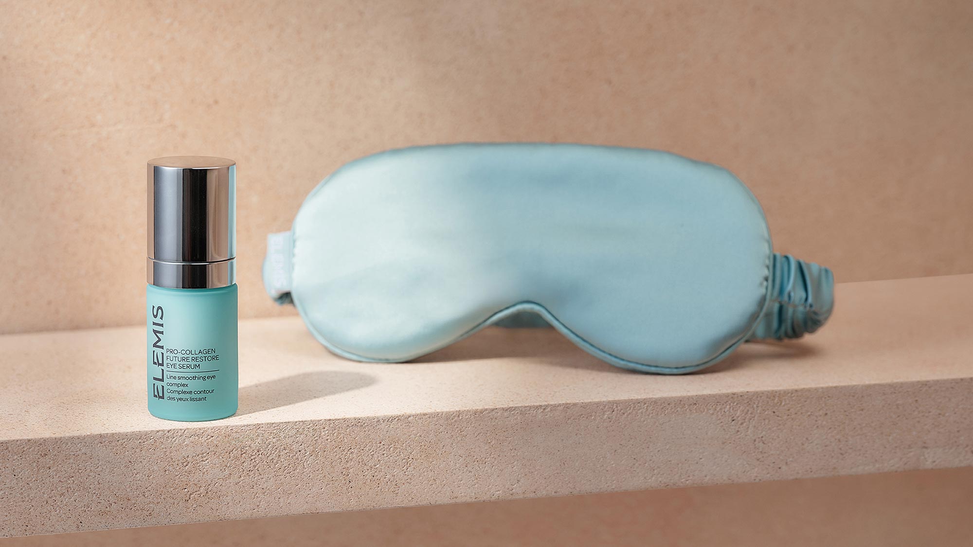 Elemis Pro-Collagen Future Restore Eye Serum in turquoise bottle next to light blue silk sleep mask on neutral background
