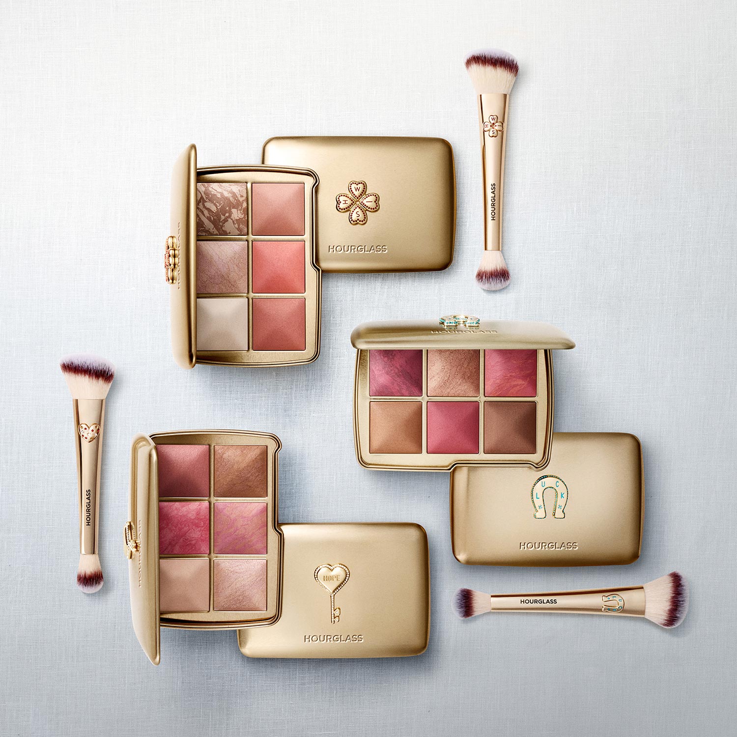 Hourglass eyeshadow palettes and makeup brushes with gold compacts in warm, neutral tones arranged on white background.