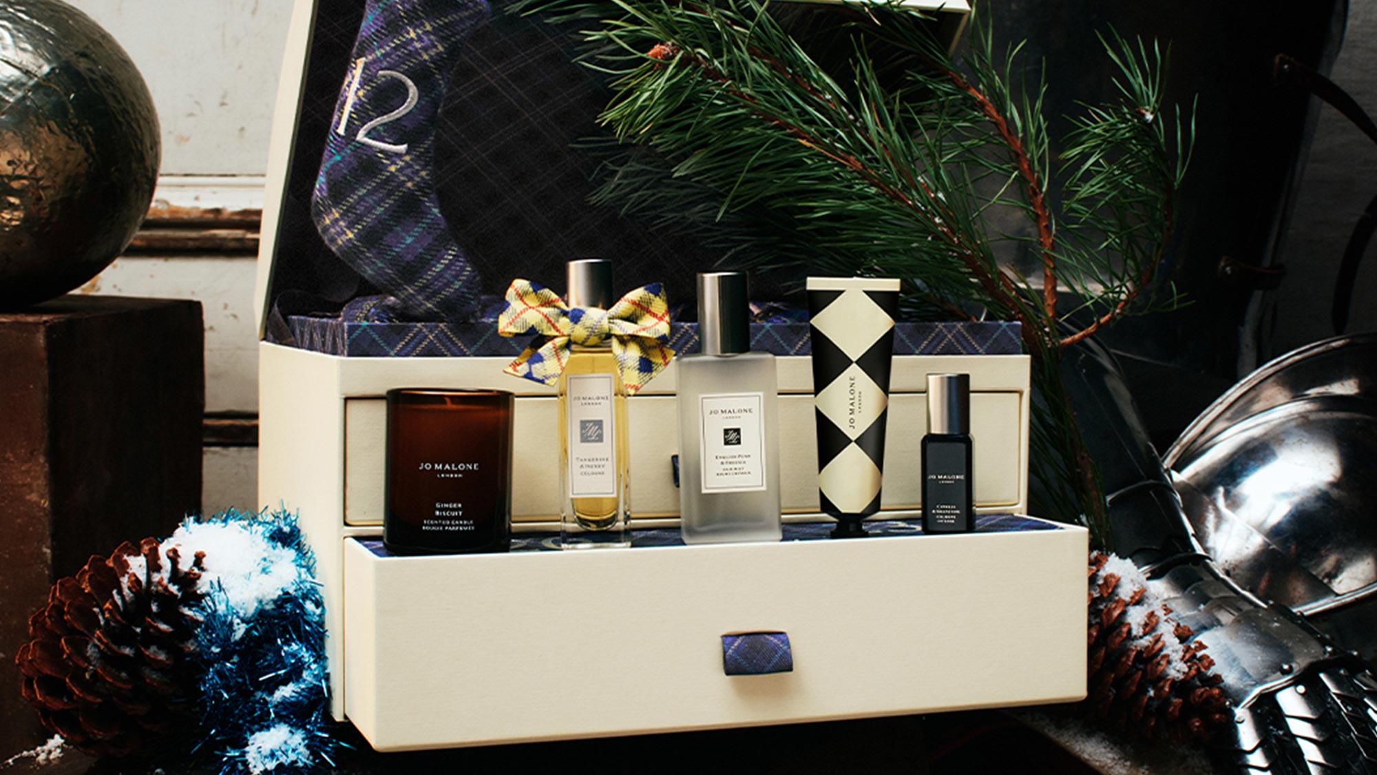 Jo Malone London fragrance collection displayed in cream gift box with blue plaid ribbon, surrounded by festive decorations and greenery
