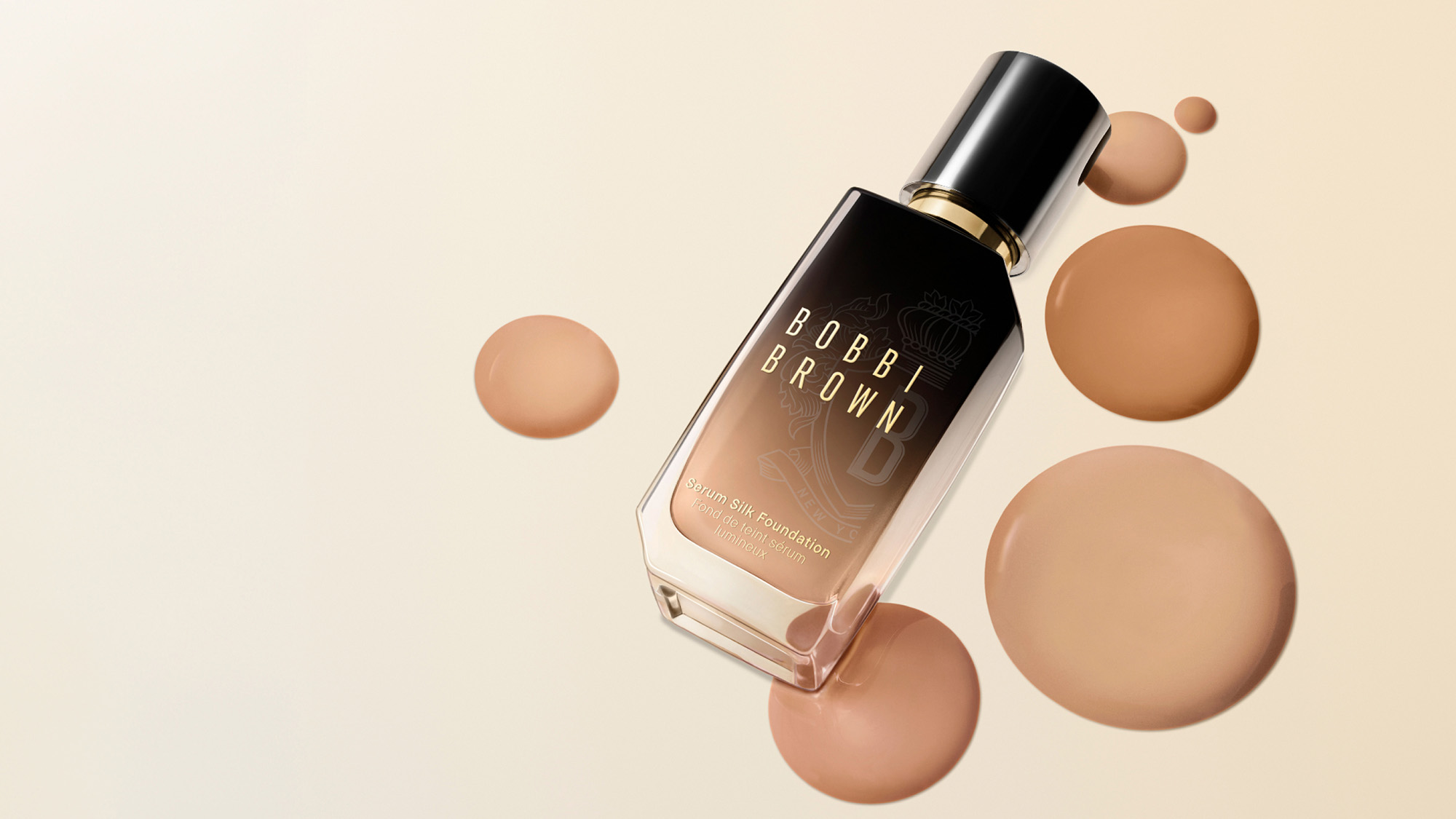 Bobbi Brown Serum Silk Foundation bottle with black cap and gold trim surrounded by beige foundation droplets on cream background.
