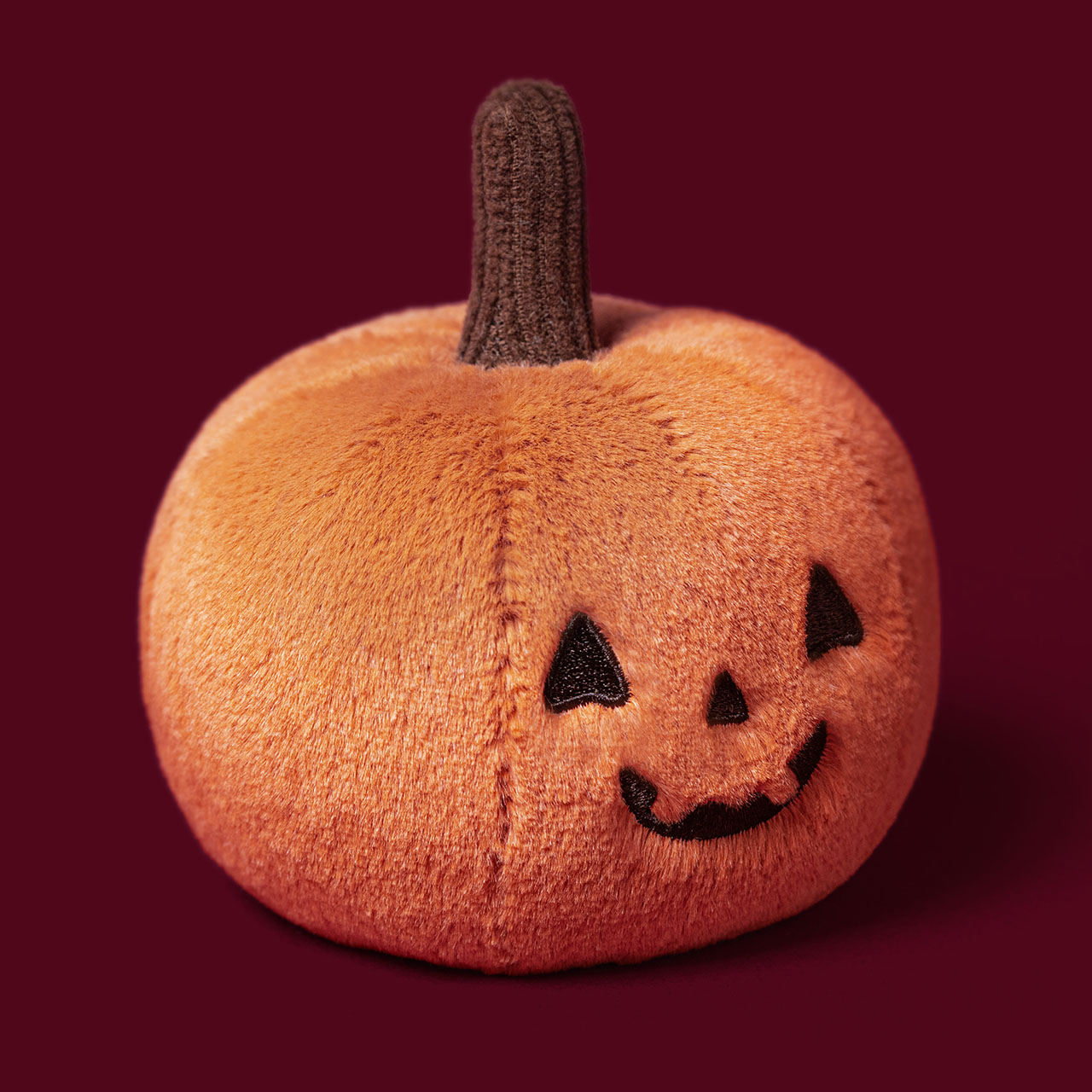Orange plush pumpkin toy with black carved jack-o'-lantern face and brown stem against deep burgundy background