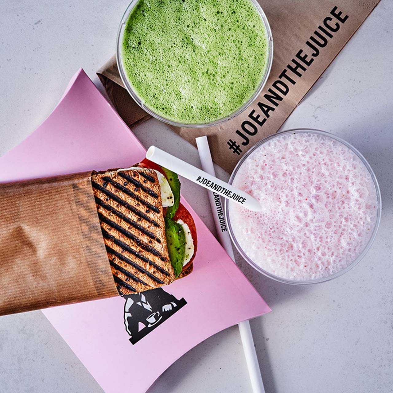 Joe & The Juice grilled sandwich with green smoothie and pink drink, featuring branded hashtag labels