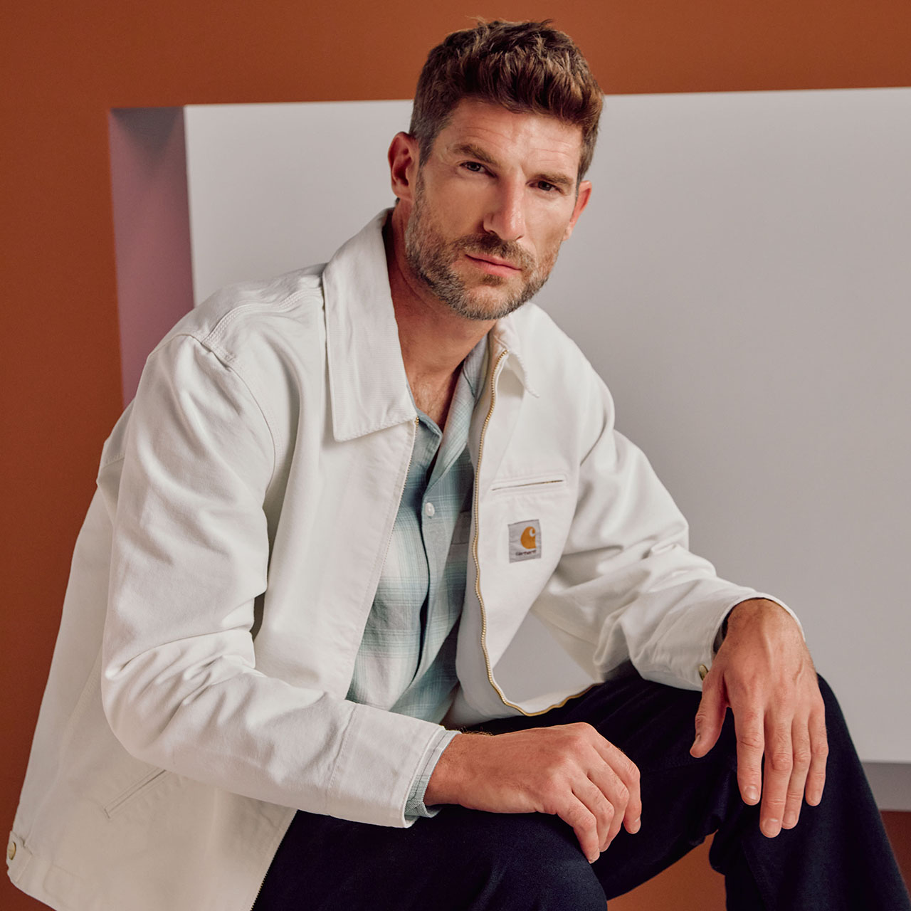 Man wearing cream utility jacket over sage green shirt and dark navy trousers, seated against orange and grey geometric background
