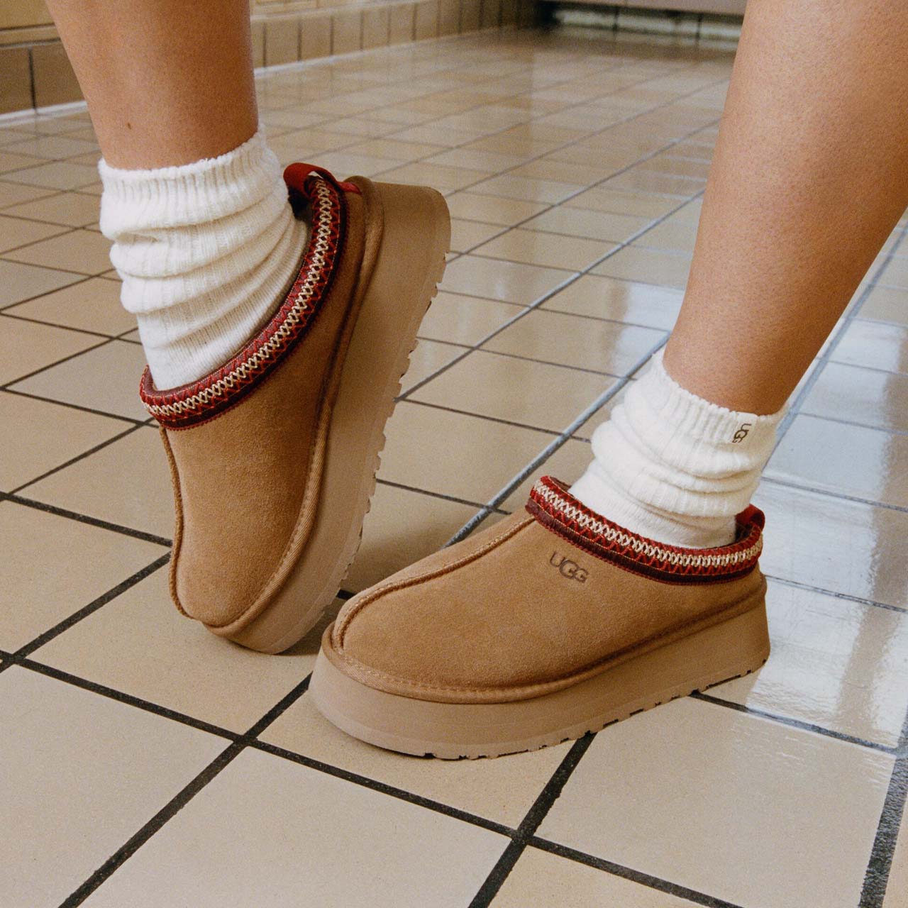 Woman wearing tan UGG boots with white ribbed socks, standing on tiled floor