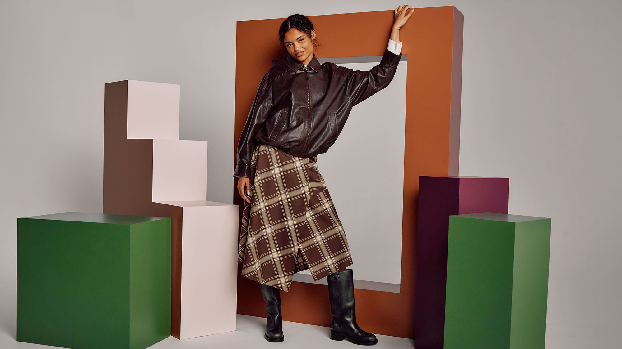 Woman wearing black leather jacket and brown tartan skirt with black boots, posing against coloured geometric backdrop