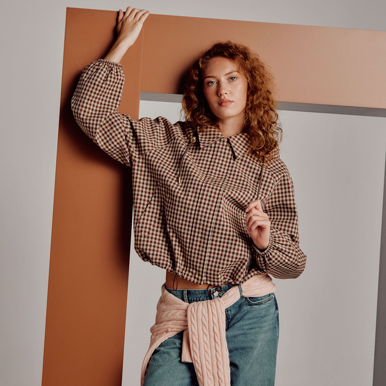 Woman with red curly hair wearing cream gingham shirt and pink cable-knit scarf, posed against geometric tan and white backdrop