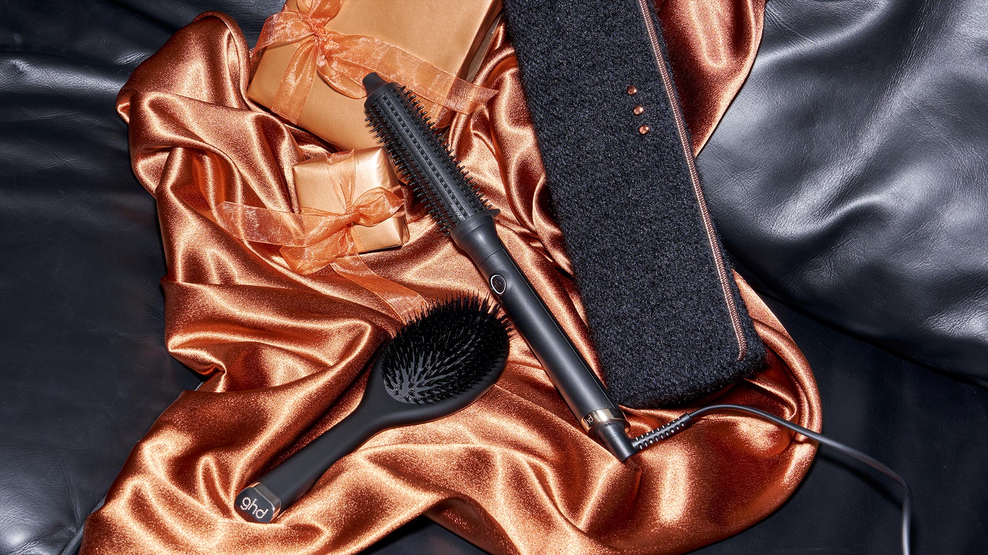 Black hair styling tool and brush on copper and bronze satin fabric with dark leather pouch against black background