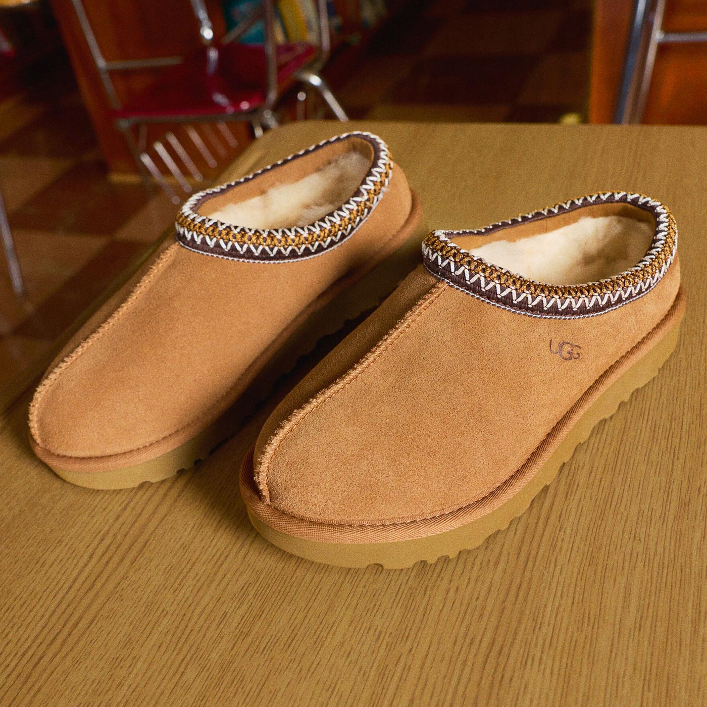 Pair of tan suede UGG slippers with white sheepskin lining and decorative whipstitched cuff on wooden surface