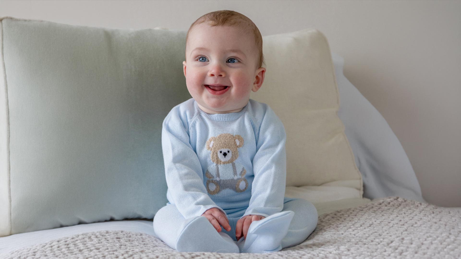 Smiling baby wearing light blue knit jumper with teddy bear design and white leggings, sitting on neutral cushions