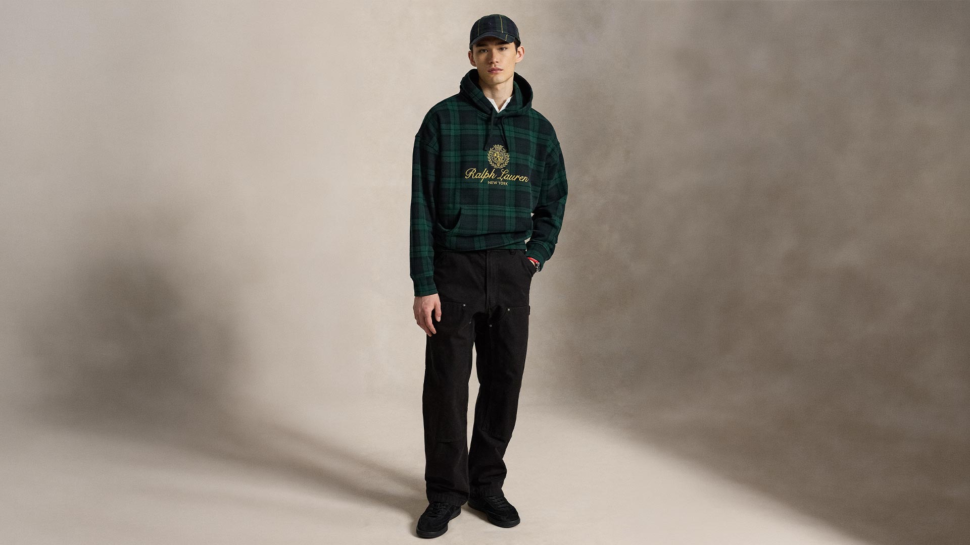 Young man wearing dark green tartan hoodie, black trousers, and dark cap against neutral background