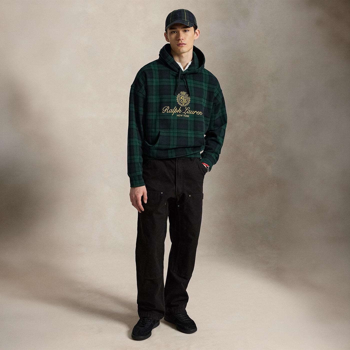 Man wearing forest green tartan hoodie with embroidered crest logo, black trousers, navy cap and black trainers