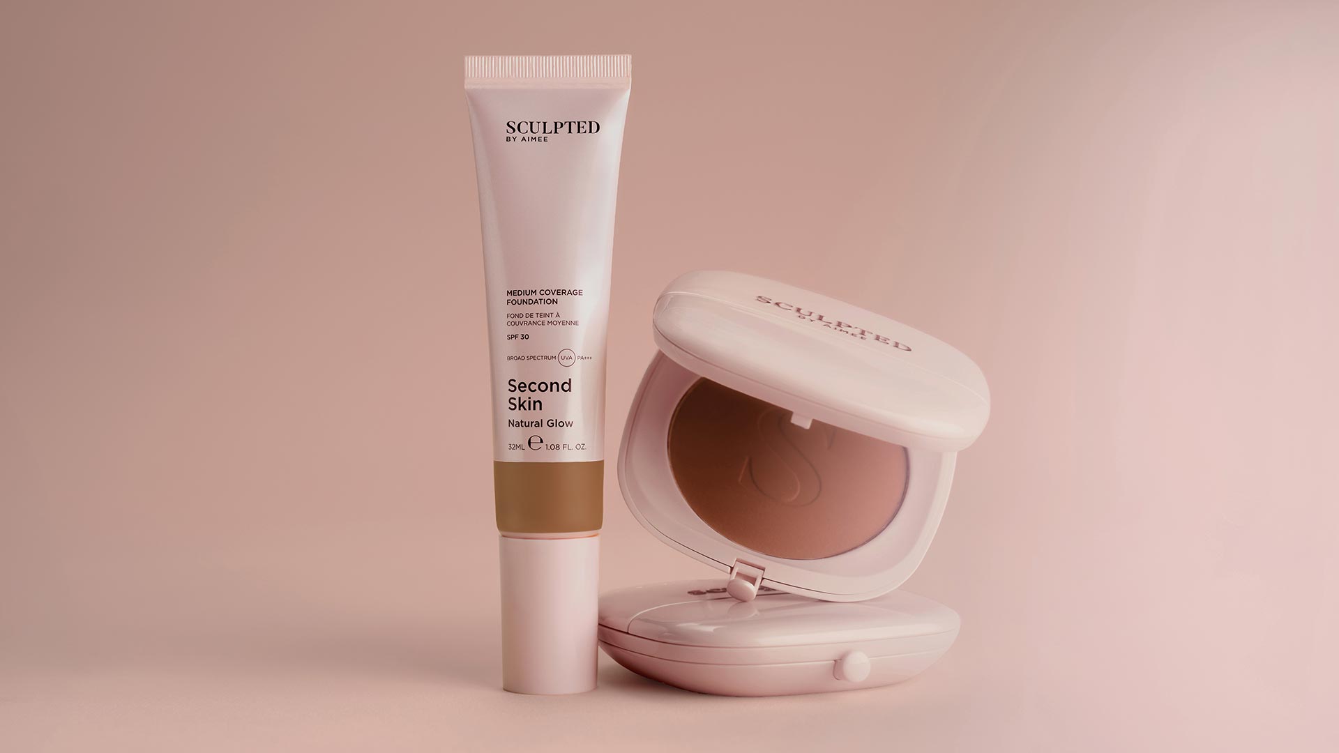 Sculpted by Aimee Second Skin foundation tube and pink powder compact with natural glow finish on blush background.