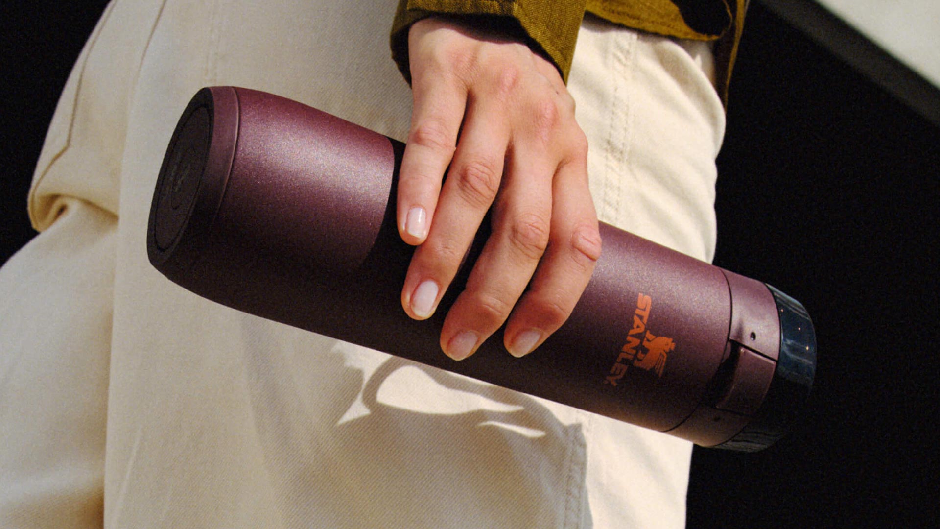Person holding burgundy Stanley thermal bottle with mustard-coloured jacket and cream trousers