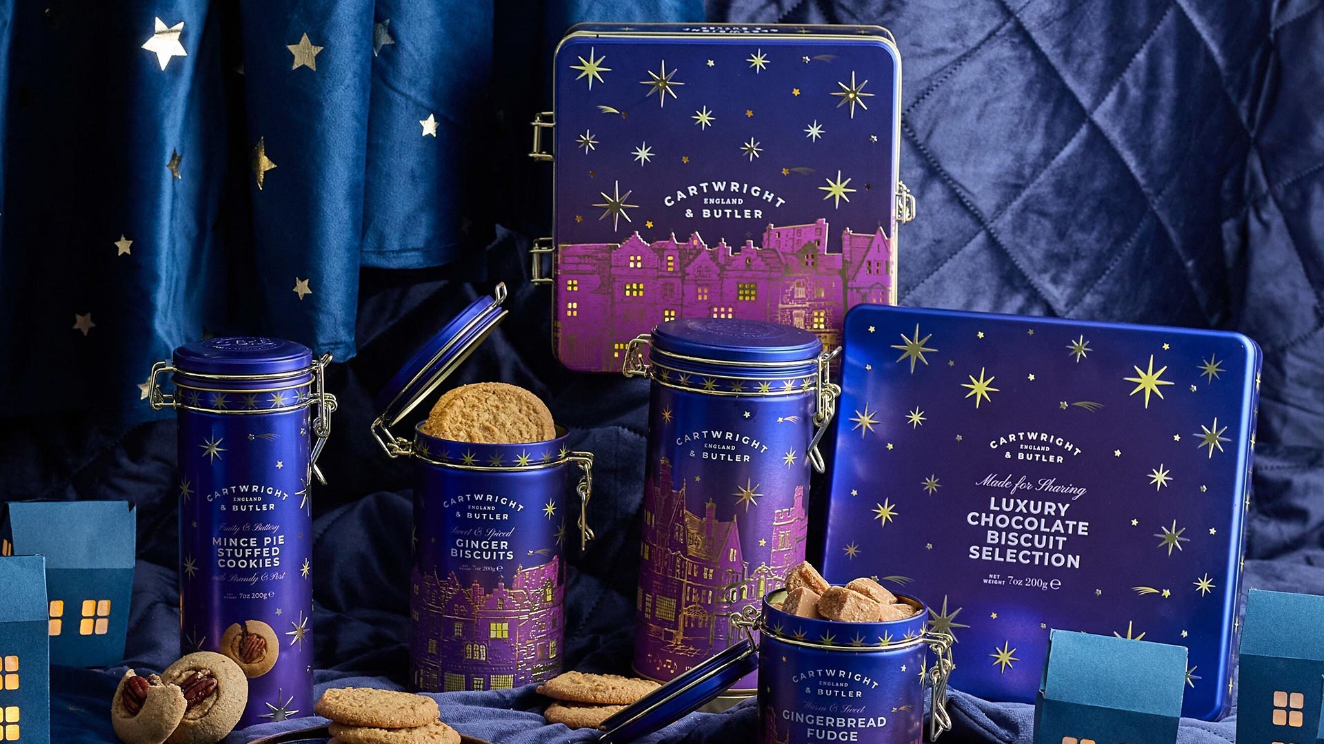 Cartwright & Butler festive gift tins and biscuits displayed with gold stars, gingerbread and chocolate treats against blue backdrop