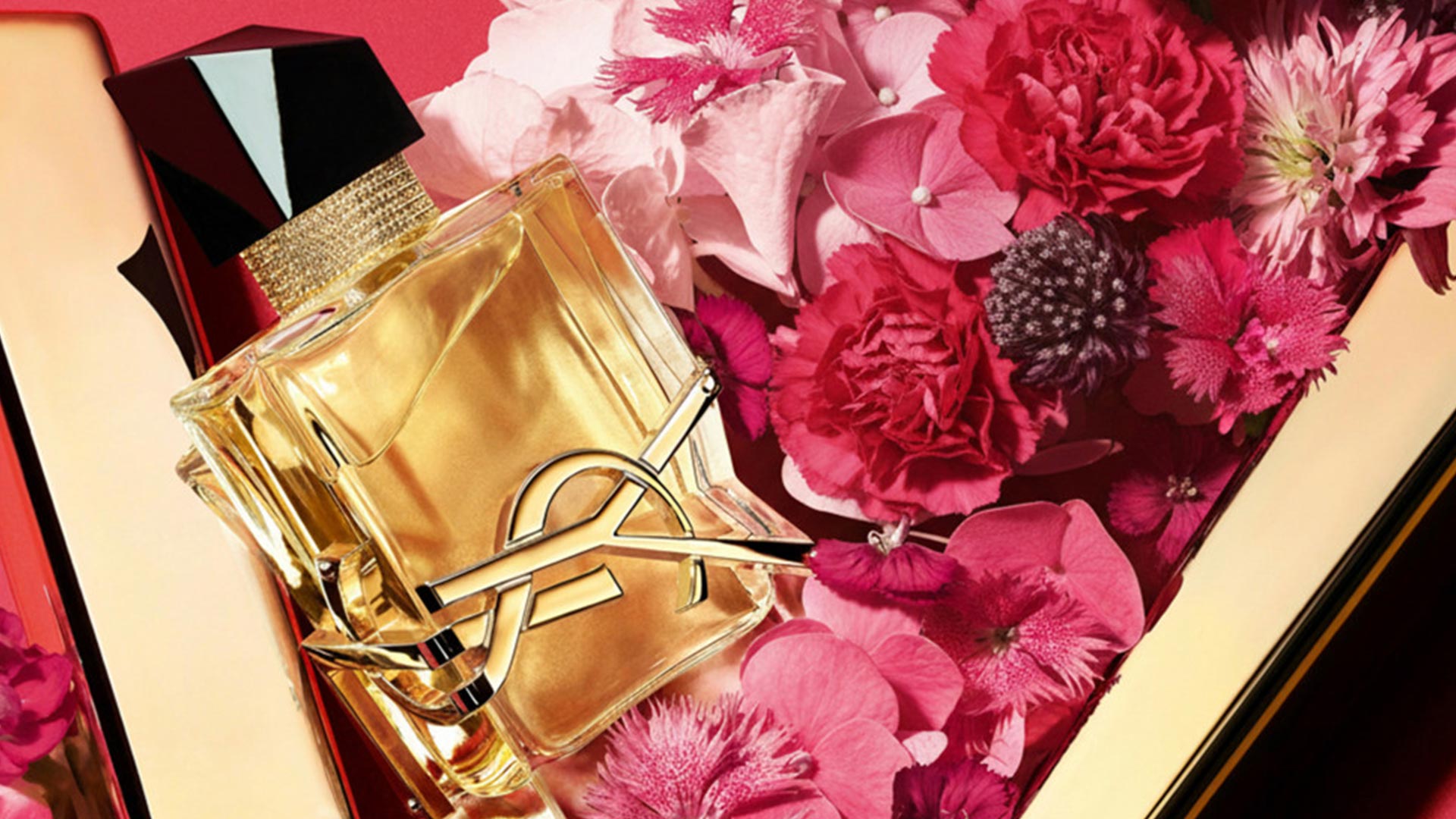 Yves Saint Laurent fragrance bottle with gold liquid and black ribbon bow, surrounded by pink and red flowers.