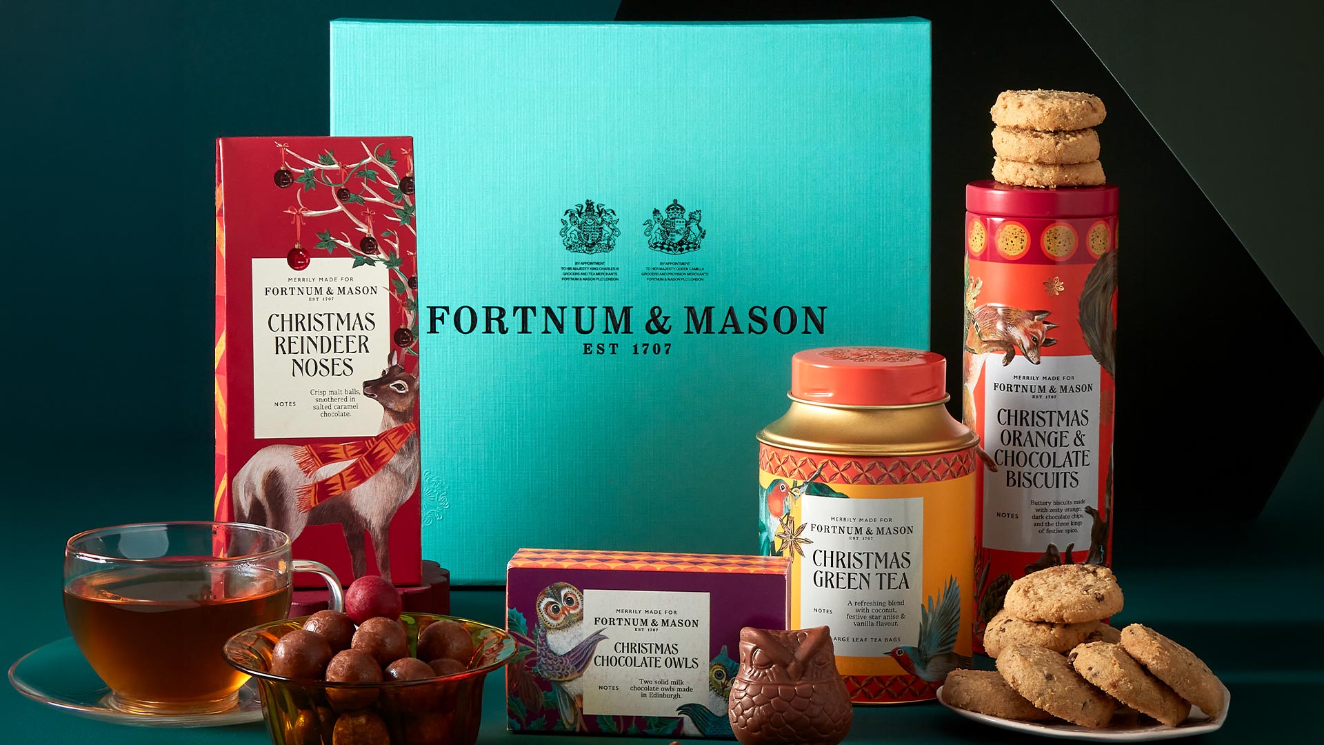 Fortnum & Mason Christmas collection featuring festive treats including reindeer noses, chocolate owls, orange and chocolate biscuits, green tea, and chocolates on turquoise background