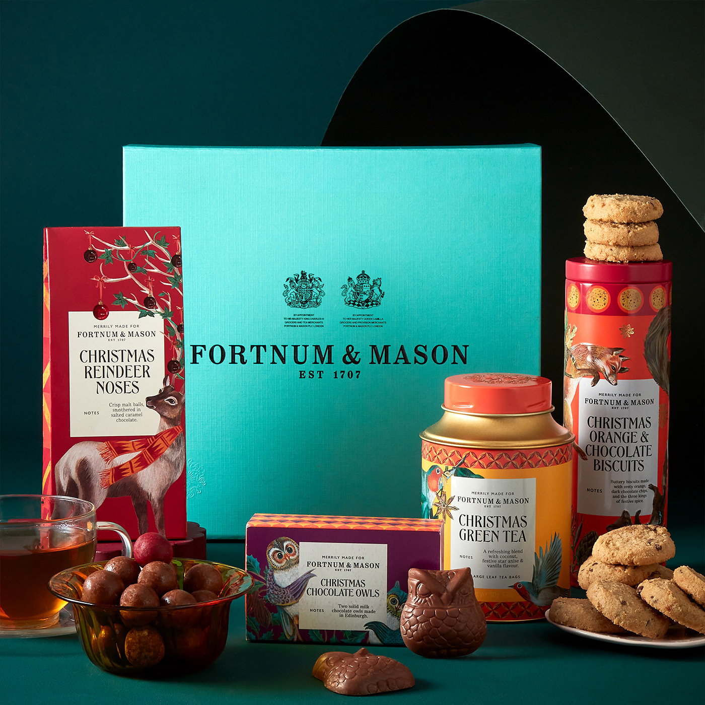 Fortnum & Mason Christmas collection featuring festive treats including reindeer noses, chocolate owls, orange and chocolate biscuits, green tea, and chocolates on turquoise background
