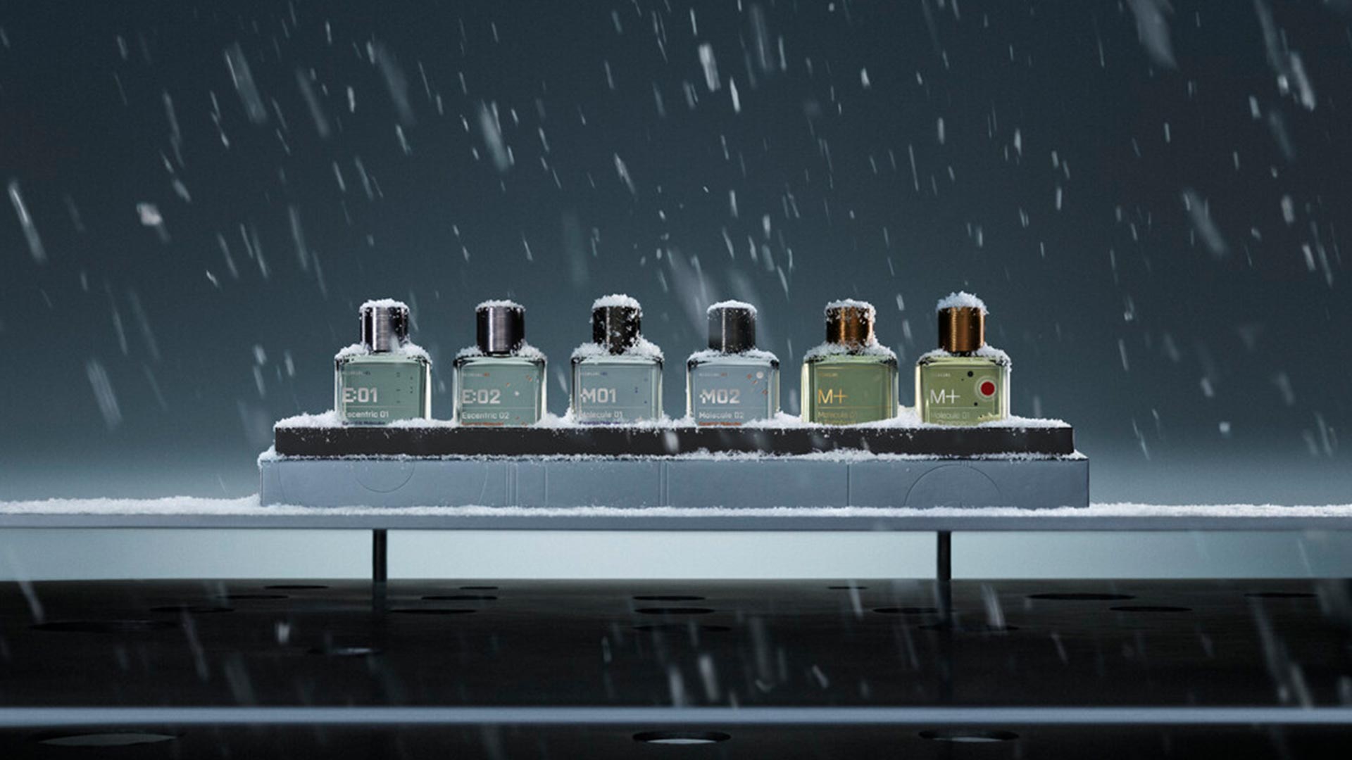 Six Escentric Molecules fragrance bottles with snow-covered caps arranged on a platform in falling snow