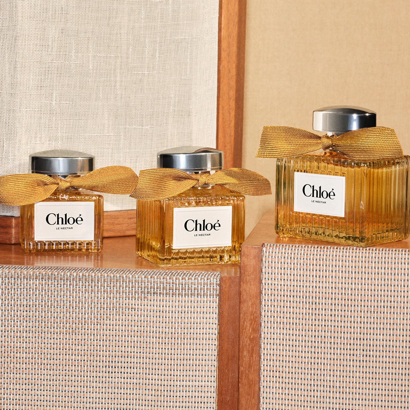 Three Chlo&eacute; Le Nectar fragrance bottles with golden bows on wooden shelf against neutral background
