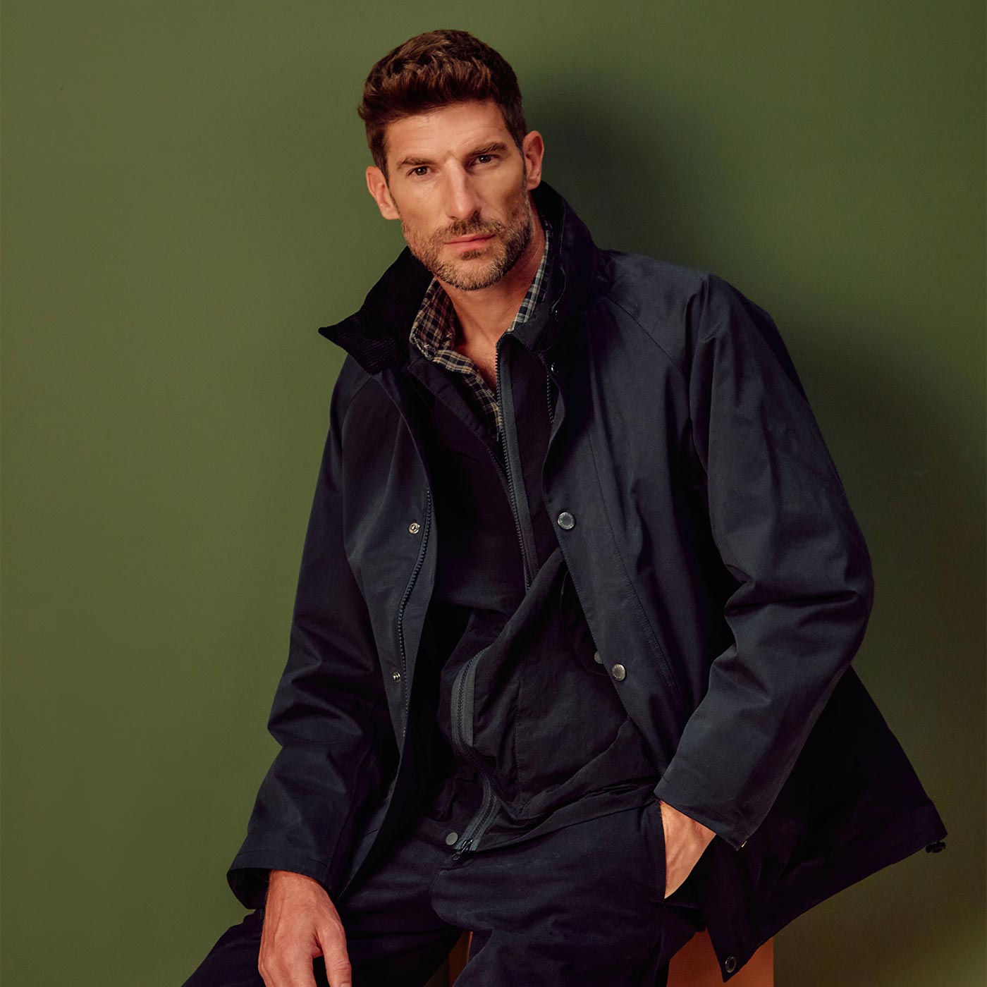 Man wearing navy jacket with checked shirt underneath against green background