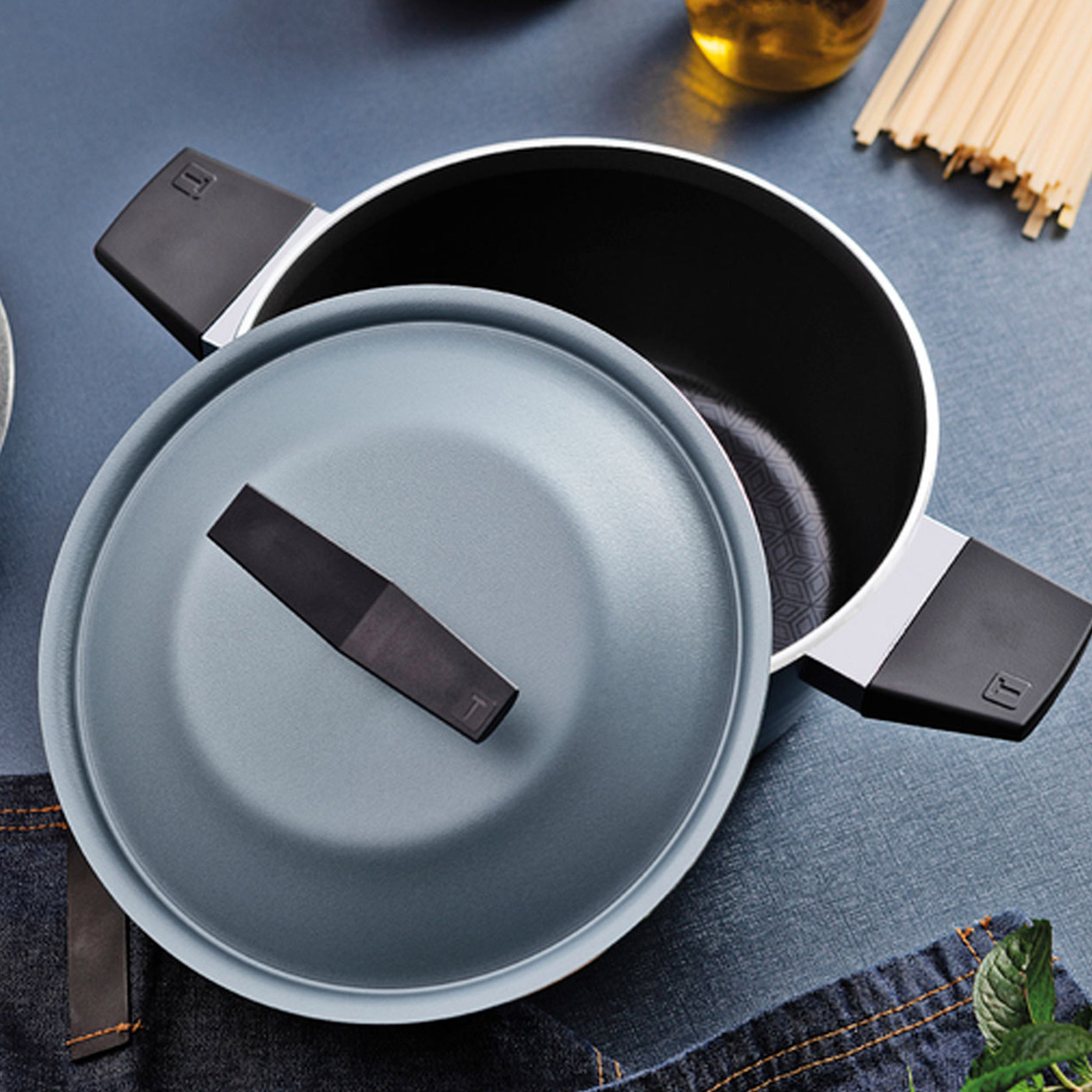 Grey cooking pot with black handles and matching lid, surrounded by kitchen utensils, oil bottle, and wooden spoons on blue surface