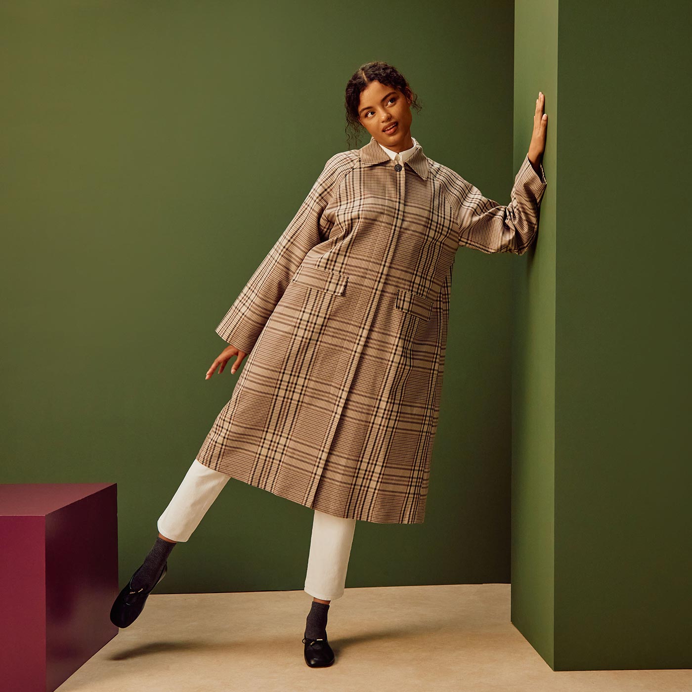 Woman wearing beige checked shirt dress with white cropped trousers and black shoes against green background
