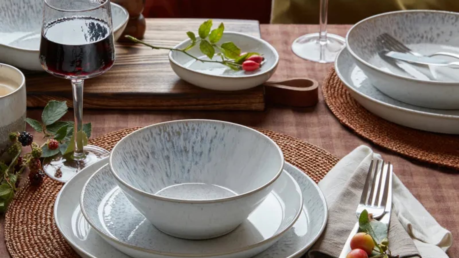 Elegant table setting with ceramic dishes, wine glasses, a candle, and decorative berries on woven mats and napkins.