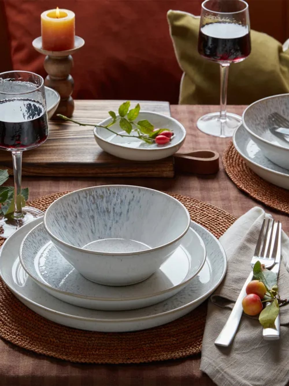 Elegant table setting with ceramic dishes, wine glasses, a candle, and decorative berries on woven mats and napkins.
