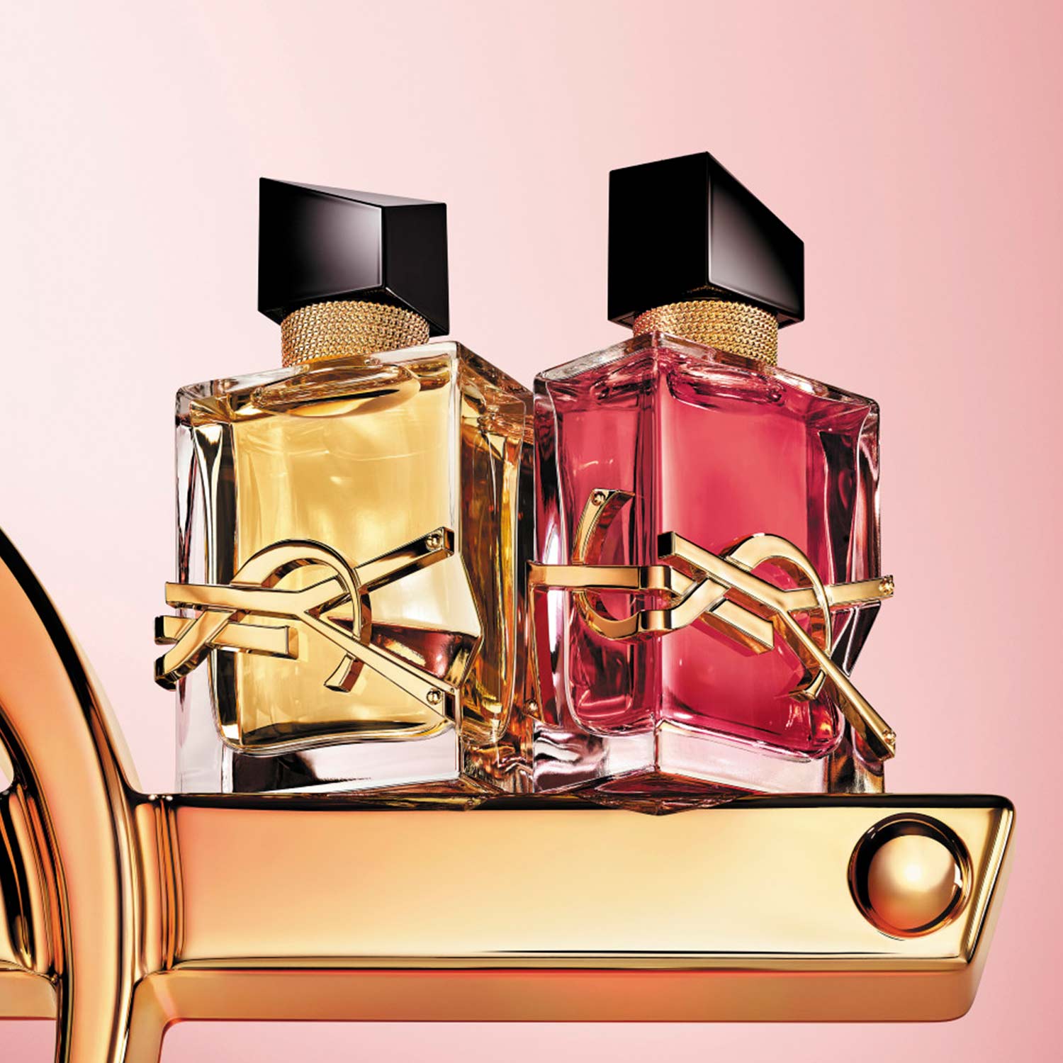 Two perfume bottles, one yellow and one pink, with gold accents, on a gold shelf against a pink background.
