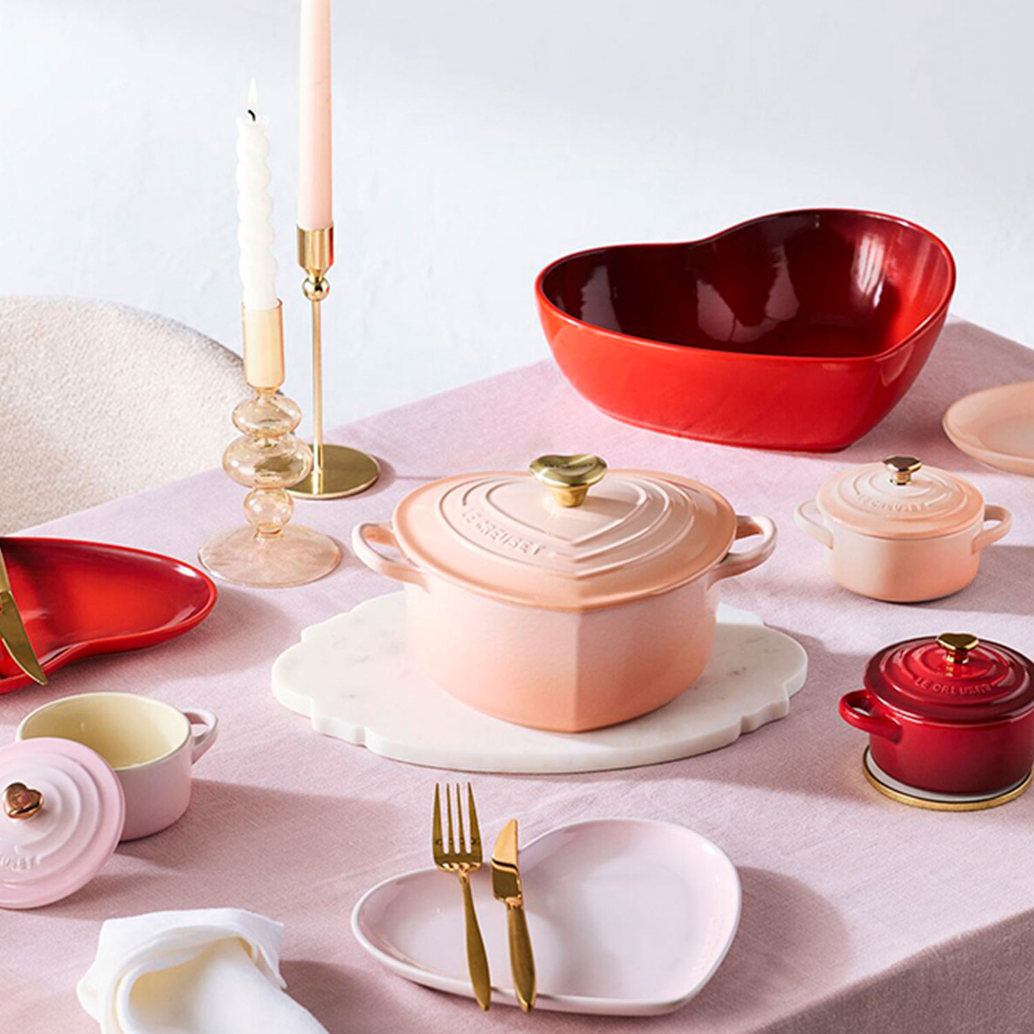 Elegant table setting with heart-shaped dishes, pastel and red tones, candles, and gold cutlery on a pink tablecloth.
