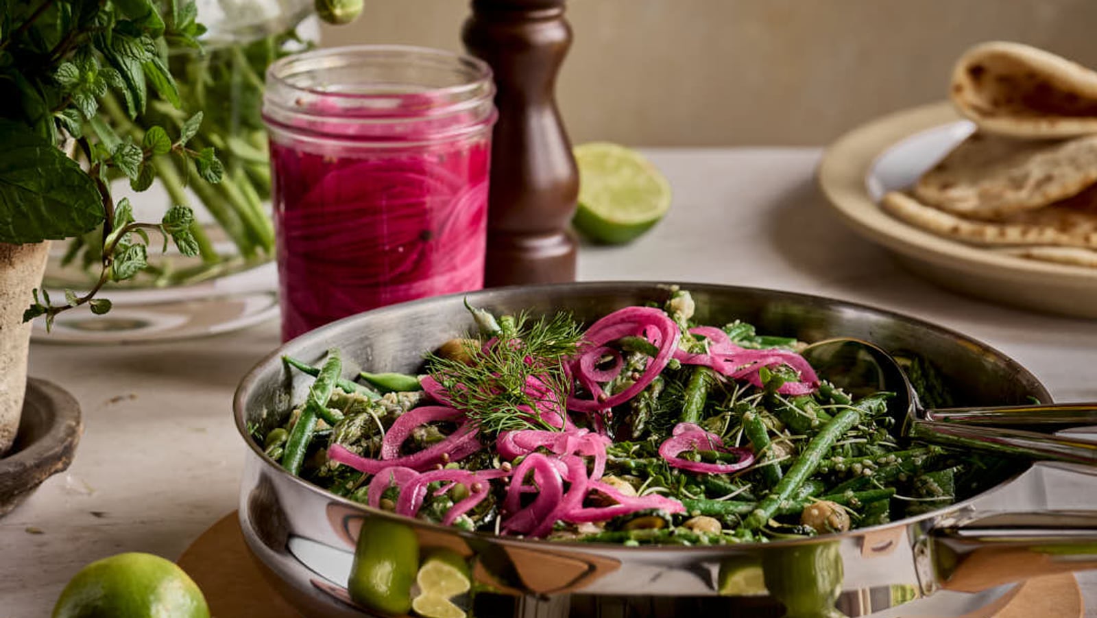 A skillet with green beans, pink pickled onions, and herbs on a table with limes, flowers, and flatbread.