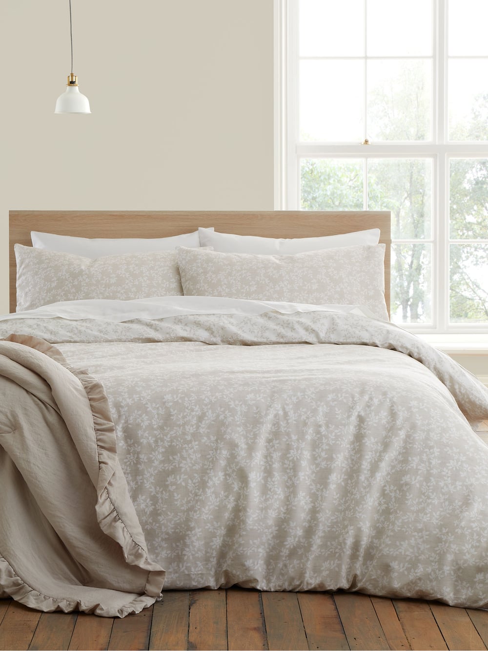 A cozy bedroom with a bed featuring blue floral bedding, framed botanical artwork, a bedside table with a plant, and a sunlit window.
