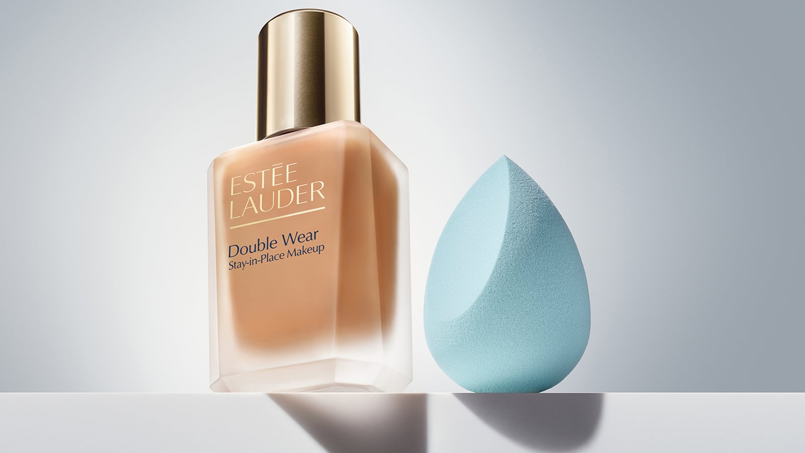 A bottle of Estée Lauder Double Wear foundation next to a light blue makeup sponge on a reflective surface.