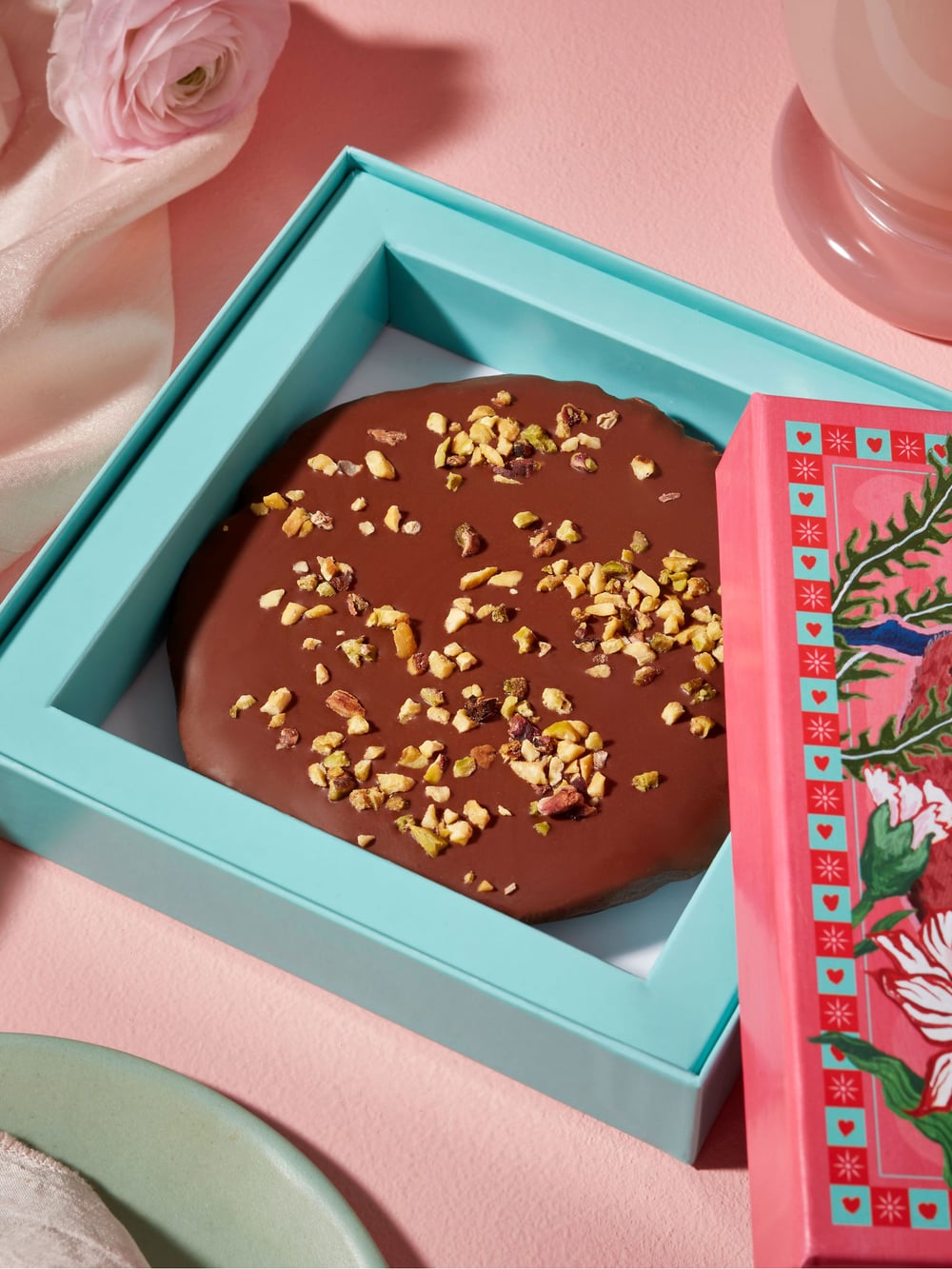 A chocolate-covered treat with nuts in a turquoise box next to a pink drink and decorative packaging.