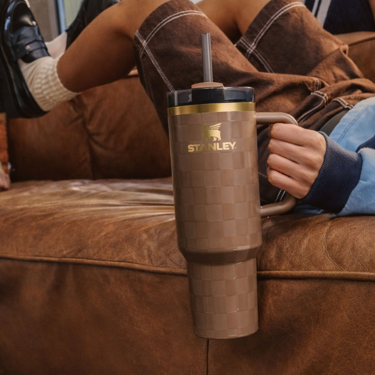 A person holding a large brown tumbler with a straw, sitting on a brown leather couch, while another person sits nearby.