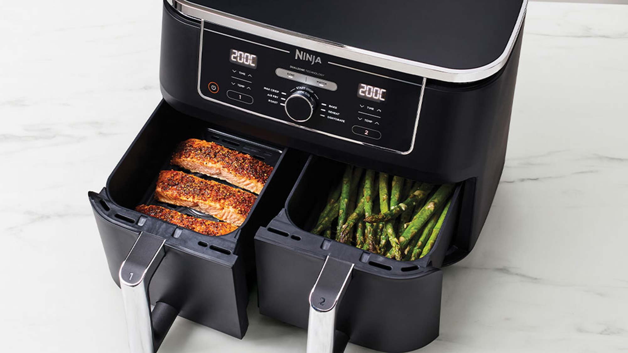Dual-compartment air fryer with cooked salmon and asparagus on a kitchen counter.