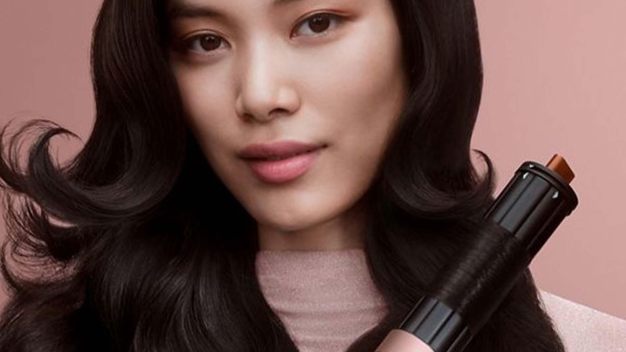 A woman with long, wavy hair holds a copper-colored hair styling tool against a pink background.