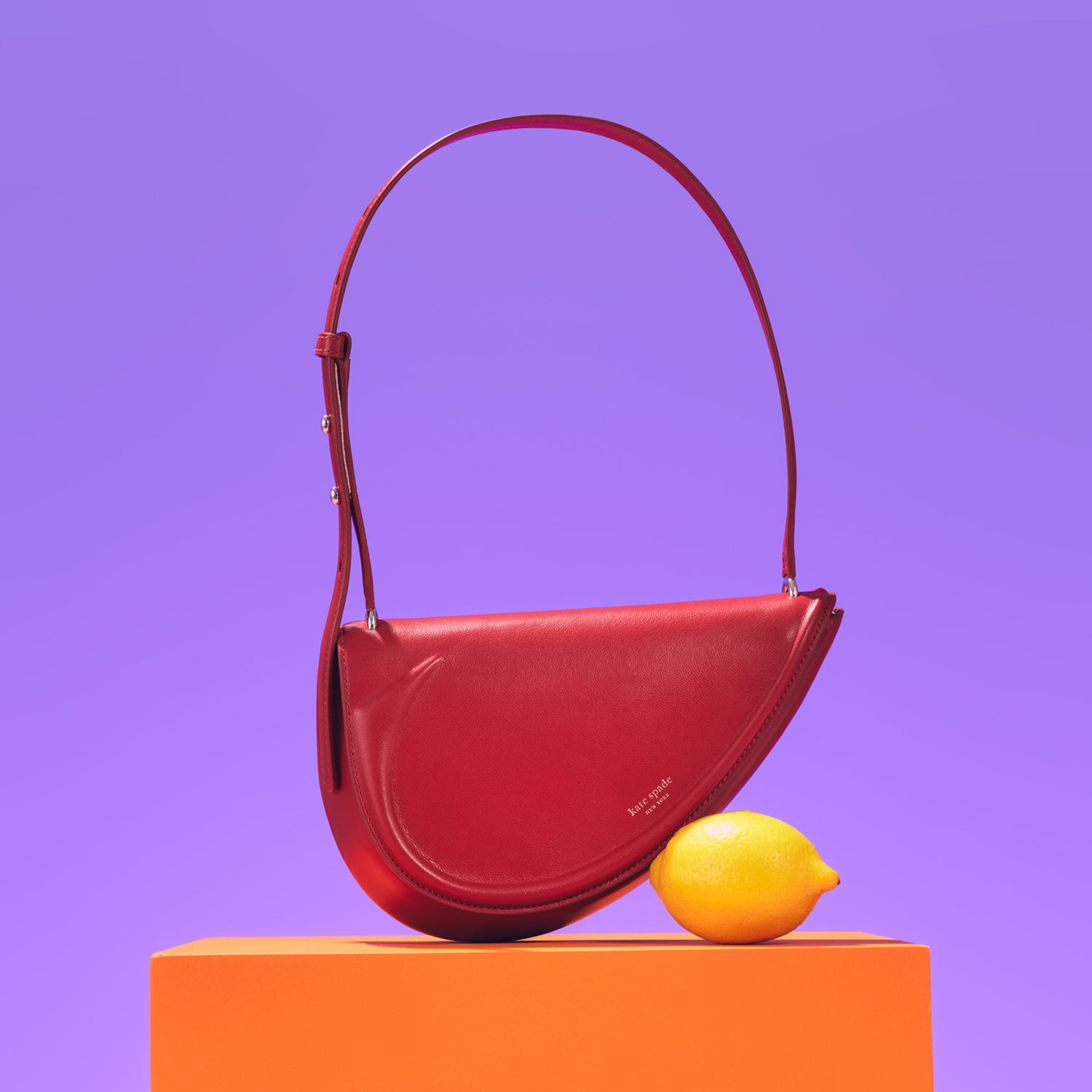 Red handbag with a curved design on an orange platform, next to a lemon, against a purple background.