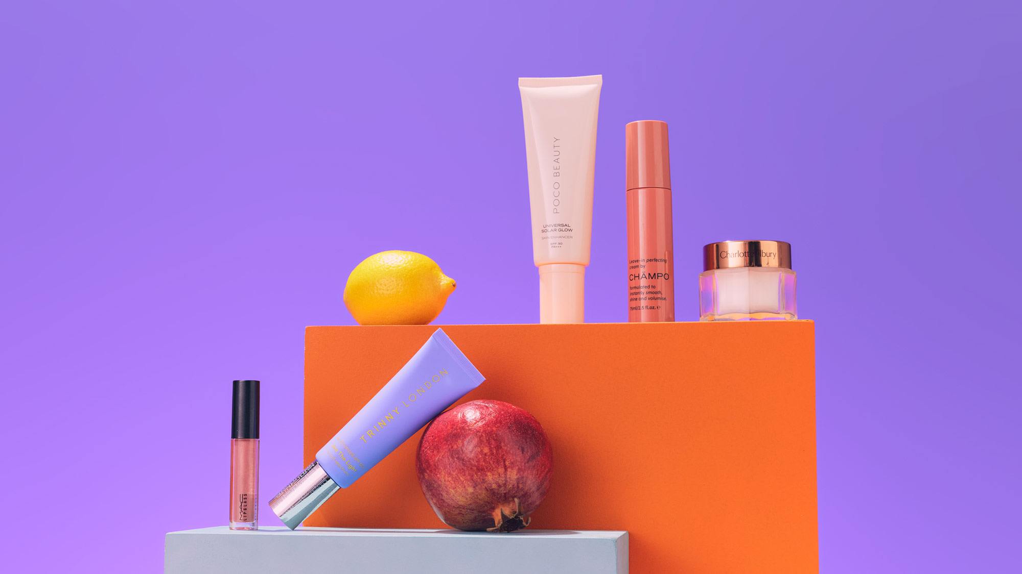 Cosmetics and fruits on colorful blocks with a purple background, featuring a lemon and pomegranate.