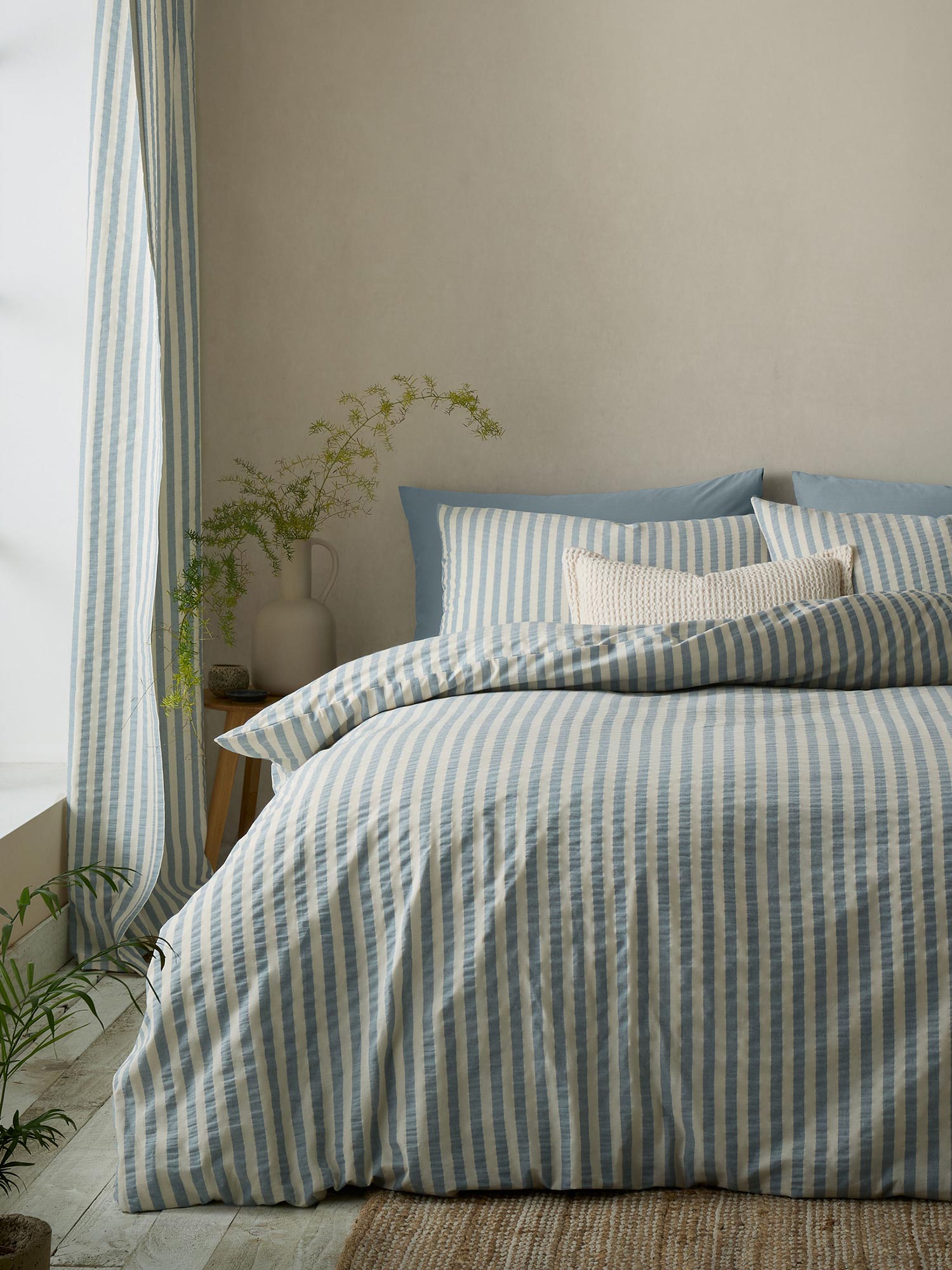 A bed with blue and white striped bedding, two pillows, and a small off-white pillow. A plant sits on a bedside table in a light room.