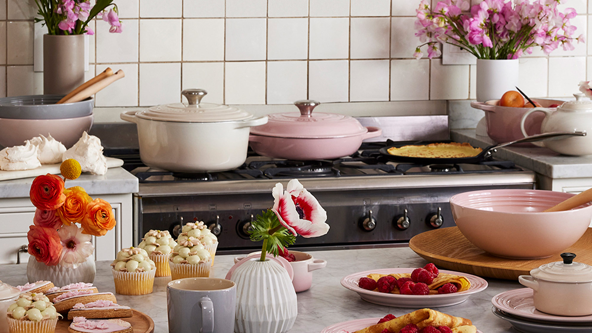 A kitchen scene with colorful flowers, cookware, and a variety of pastries on the countertop.