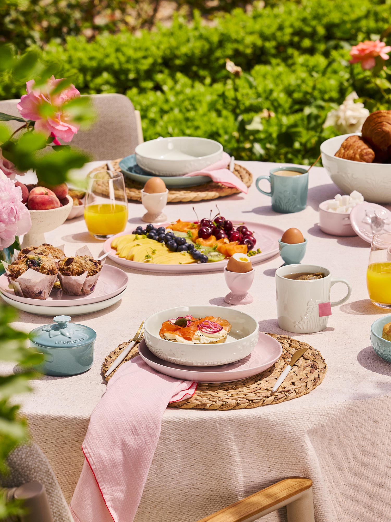 A garden table set with a colorful breakfast spread, including eggs, fruit, pastries, and drinks, surrounded by greenery and flowers.
