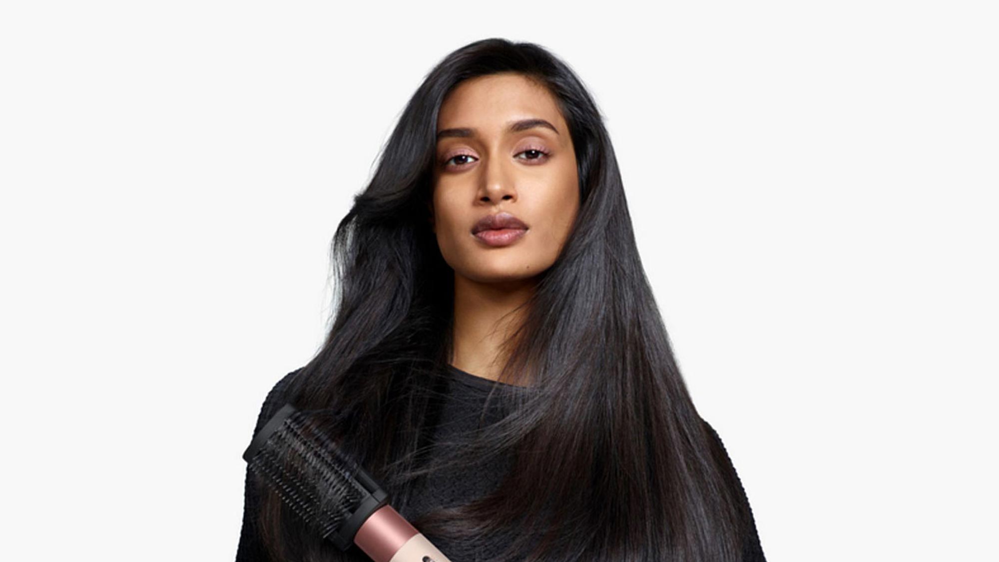 A person with long, straight dark hair holds a hair dryer brush, wearing a black top against a plain background.