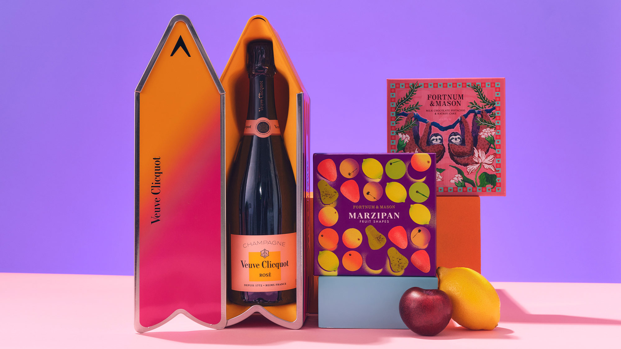 A bottle of Veuve Clicquot champagne in a gift box, surrounded by colorful marzipan packaging and fruit on a purple background.