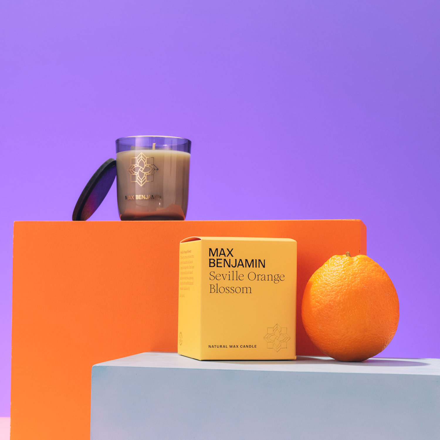 A scented candle, orange box, and orange are arranged on colorful blocks against a purple background.