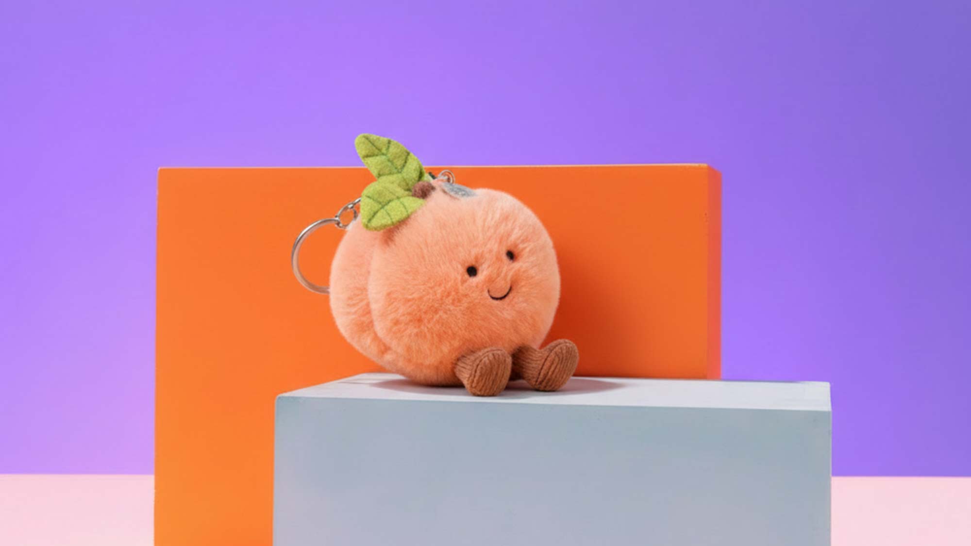 Two smiling plush clover toys, one as a keychain, on a blue block with a pomegranate, set against a purple background.