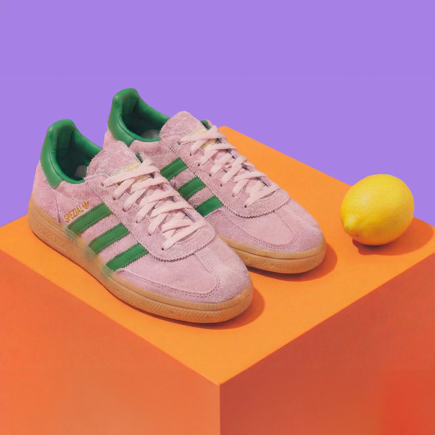 Pink sneakers with green stripes on an orange surface, next to a lemon, against a purple background.