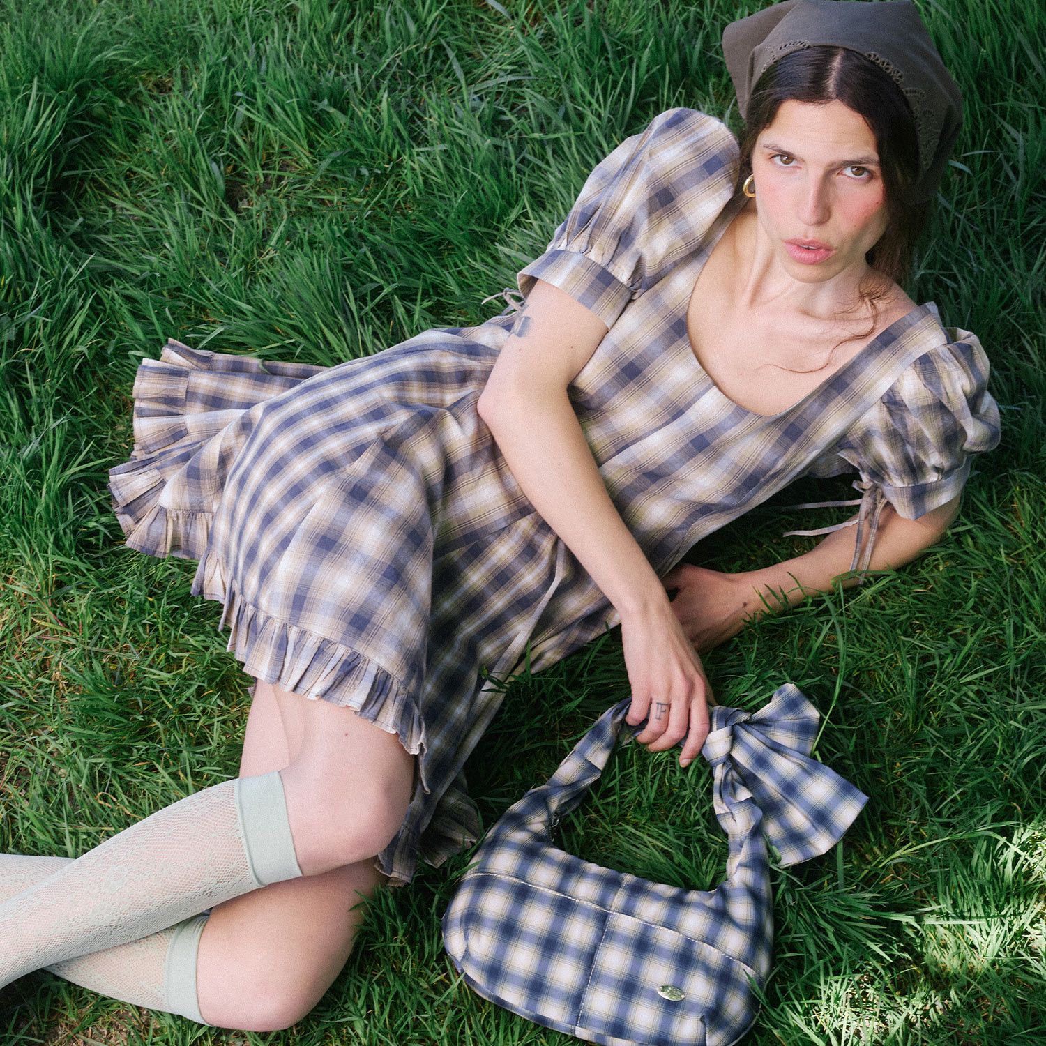 Woman wearing blue and cream plaid dress with ruffled hem, beige socks, and dark cap lying on grass
